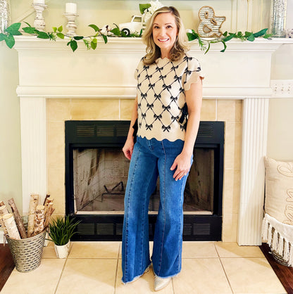 A woman stands in front of a fireplace wearing a cream short-sleeve top with a black bow pattern and blue wide-leg jeans.