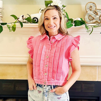 A woman wearing a pink corduroy button-up shirt with ruffled sleeves, standing in front of a fireplace mantel decorated with greenery and home decor.