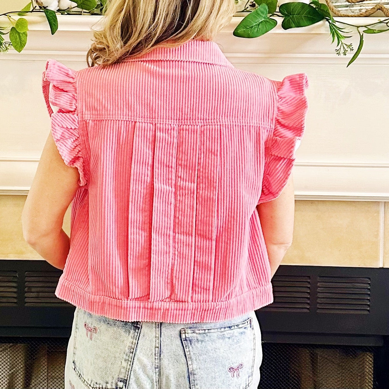 A woman viewed from behind wearing a pink and white vertically striped ruffle-sleeve crop top, standing in front of a fireplace.