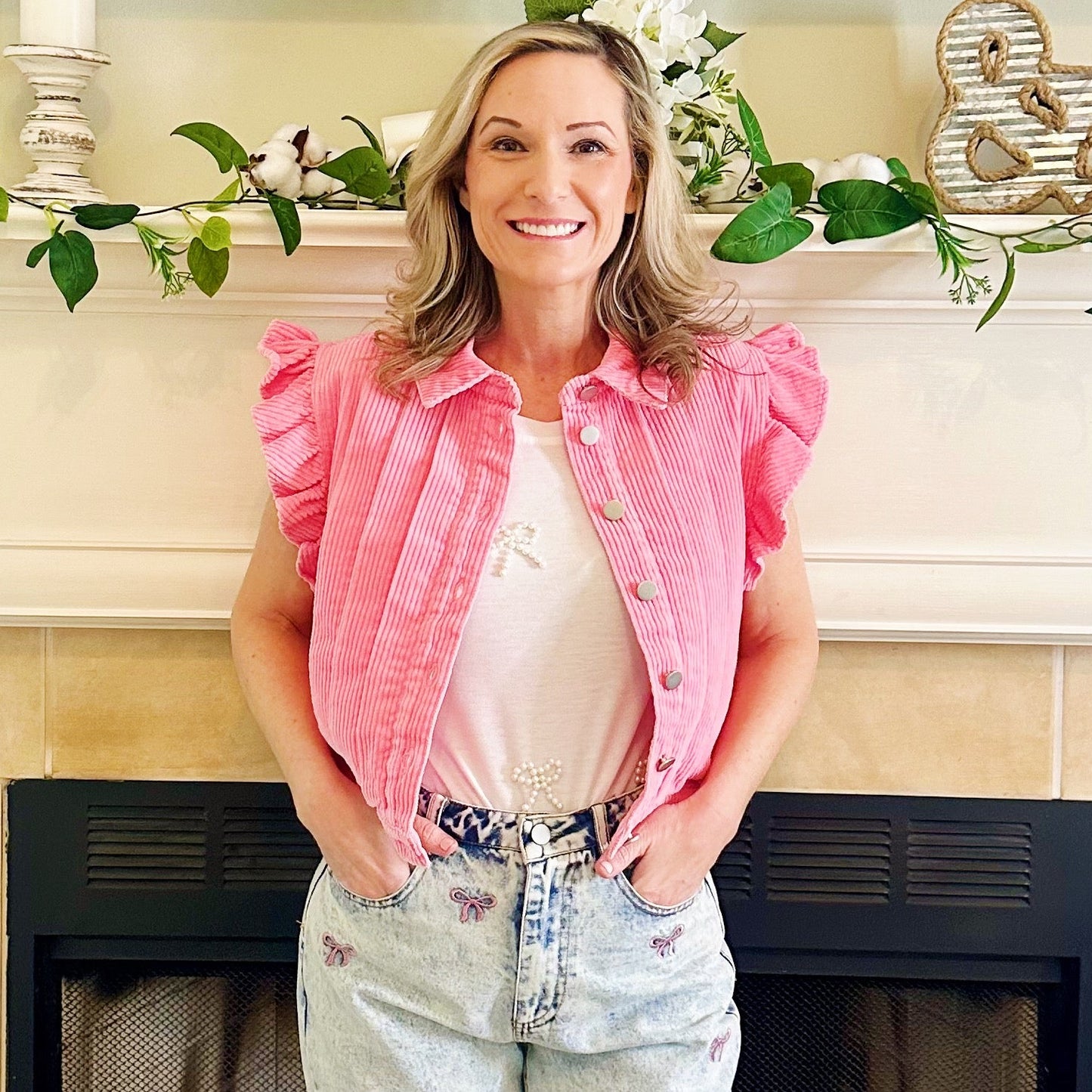 A woman stands in front of a fireplace wearing a pink corduroy cropped jacket with ruffled sleeves, a white top, and light-wash distressed jeans.