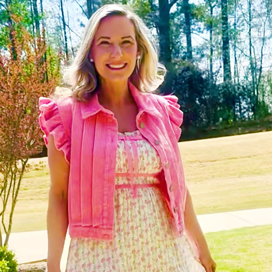 A woman stands outdoors on a paved path wearing a light pink and white floral midi dress with a matching pink ruffled cropped jacket.