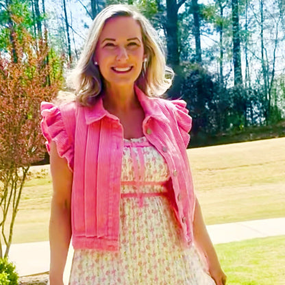 A woman stands outdoors on a paved path wearing a light pink and white floral midi dress with a matching pink ruffled cropped jacket.
