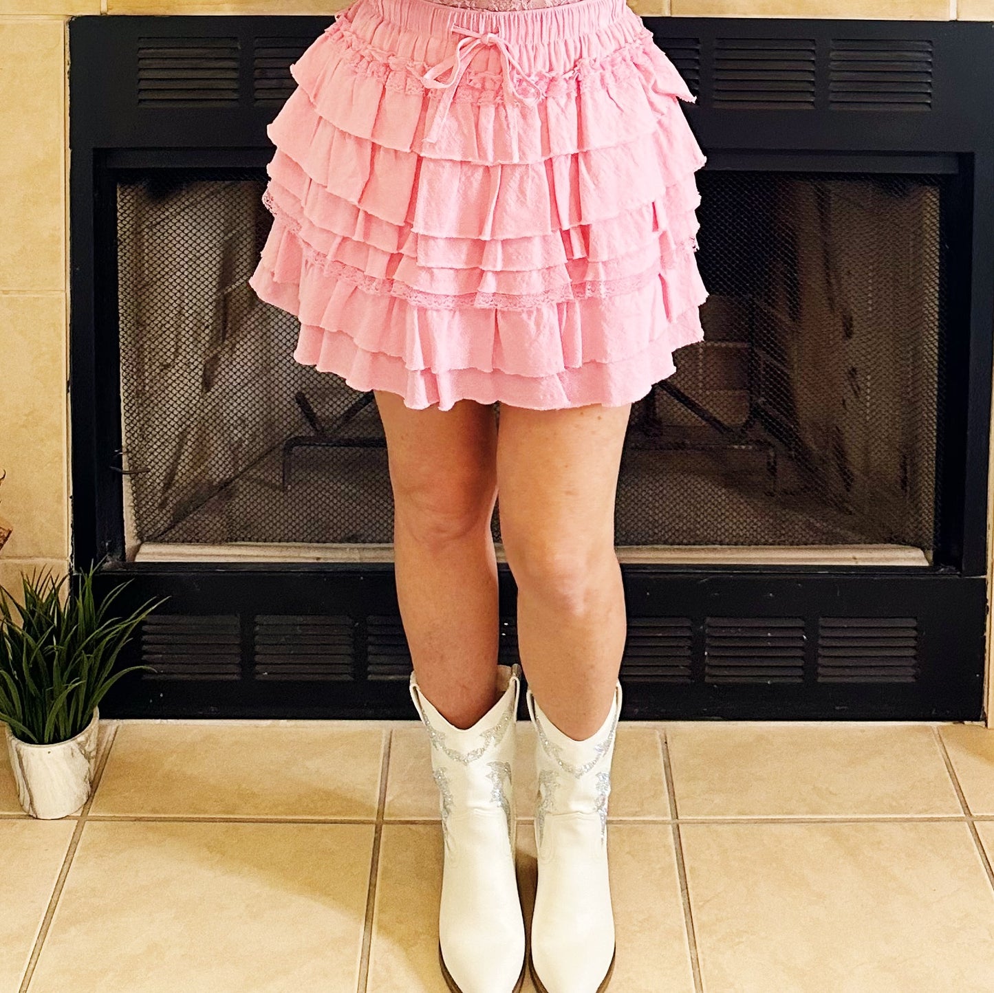 A person wearing a light pink tiered ruffle mini skirt and white western-style cowboy boots, standing in front of a black fireplace on a beige tiled floor.