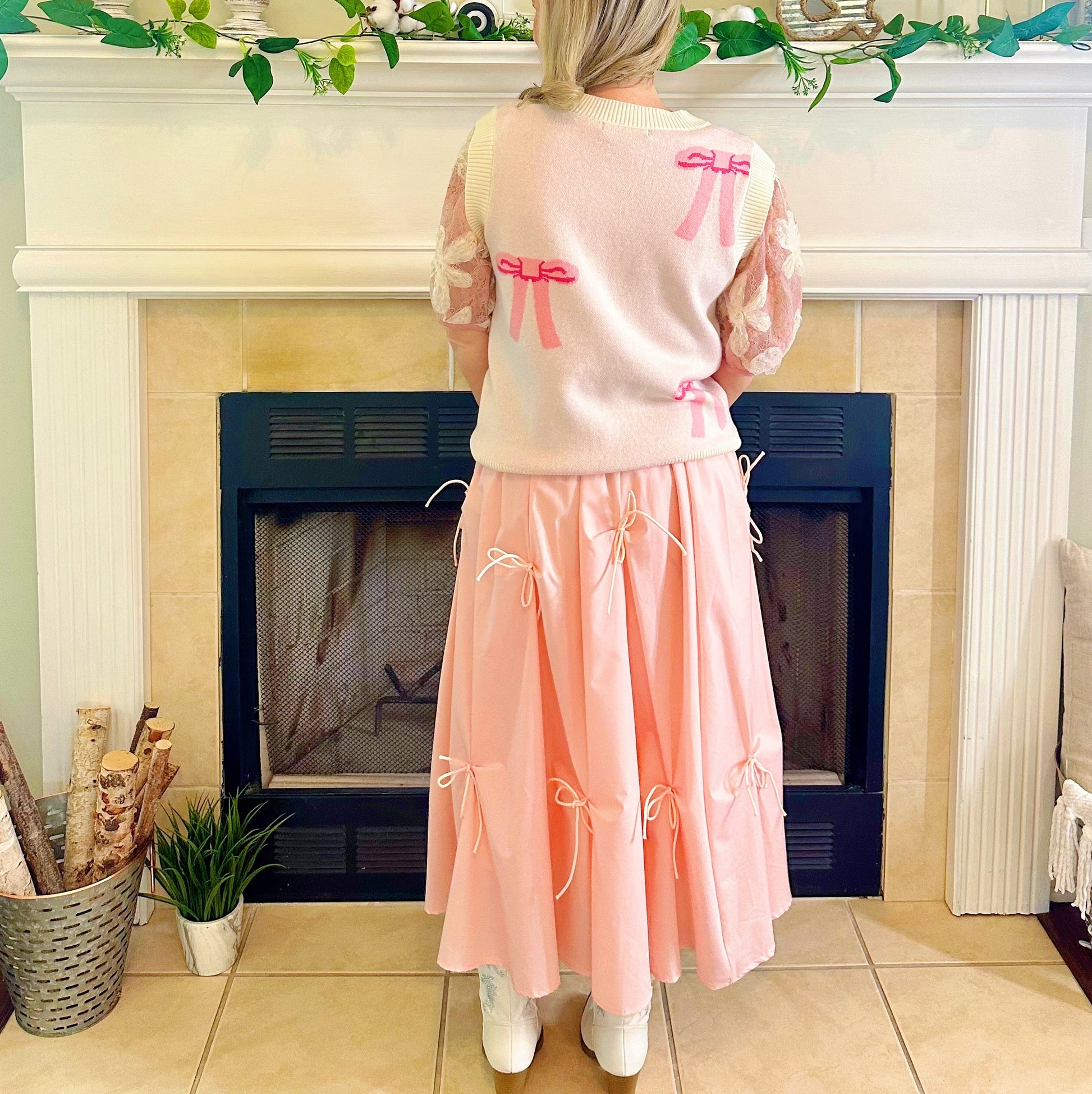 A woman stands with her back to the camera in front of a fireplace, wearing a light pink sweater with pink bow details on the back and a matching light pink pleated skirt with bow accents. The setting is a cozy, decorated indoor room with a fireplace mantel and tiled floor.