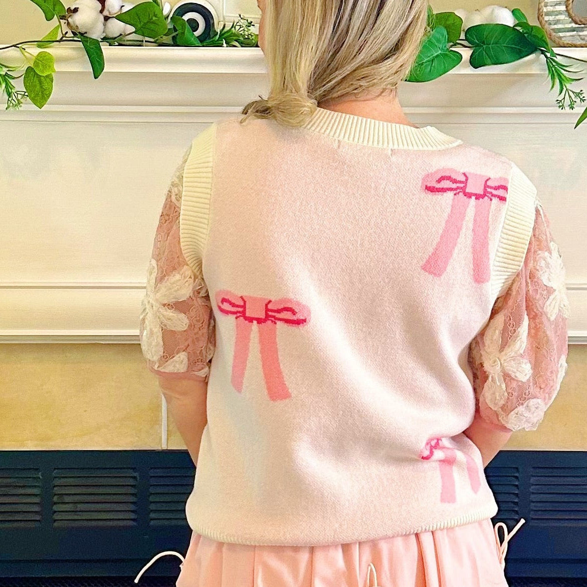 A woman stands with her back to the camera in front of a fireplace, wearing a light pink fuzzy sweater with pink bow details and a long light pink skirt with tie details.