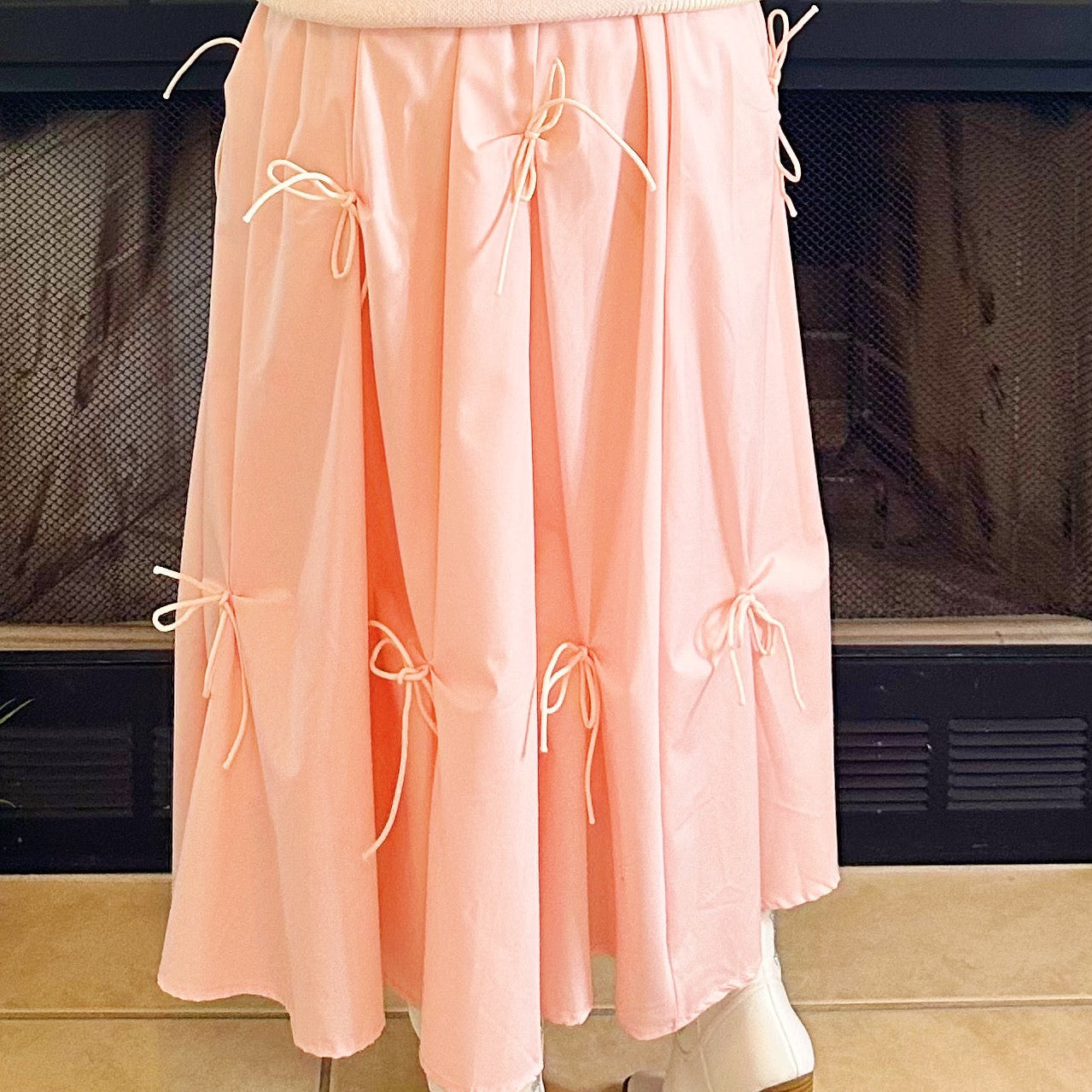 A person wearing a light pink pleated midi skirt with multiple cream-colored tie details, paired with white ankle boots, standing in front of a dark fireplace.