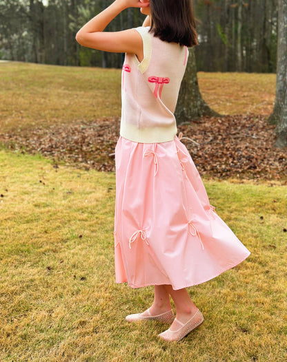 A woman standing outdoors on grass wearing a sleeveless cream knit vest with pink ribbon details and a light pink midi skirt with matching pink ribbon ties, paired with beige flats.