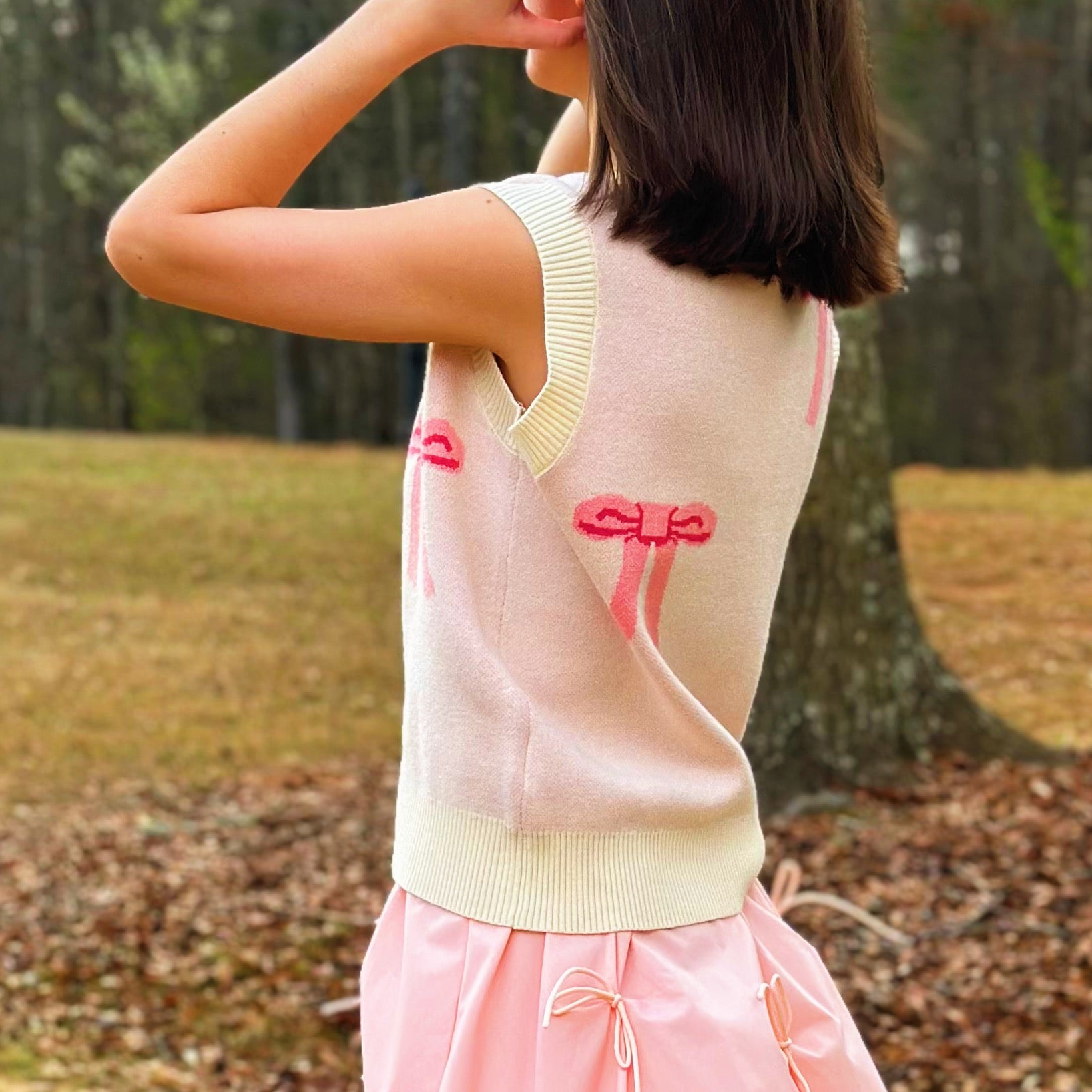 A woman wearing a pale pink sleeveless knit top with cream ribbed trim and bright pink bow details, photographed outdoors in a wooded area.