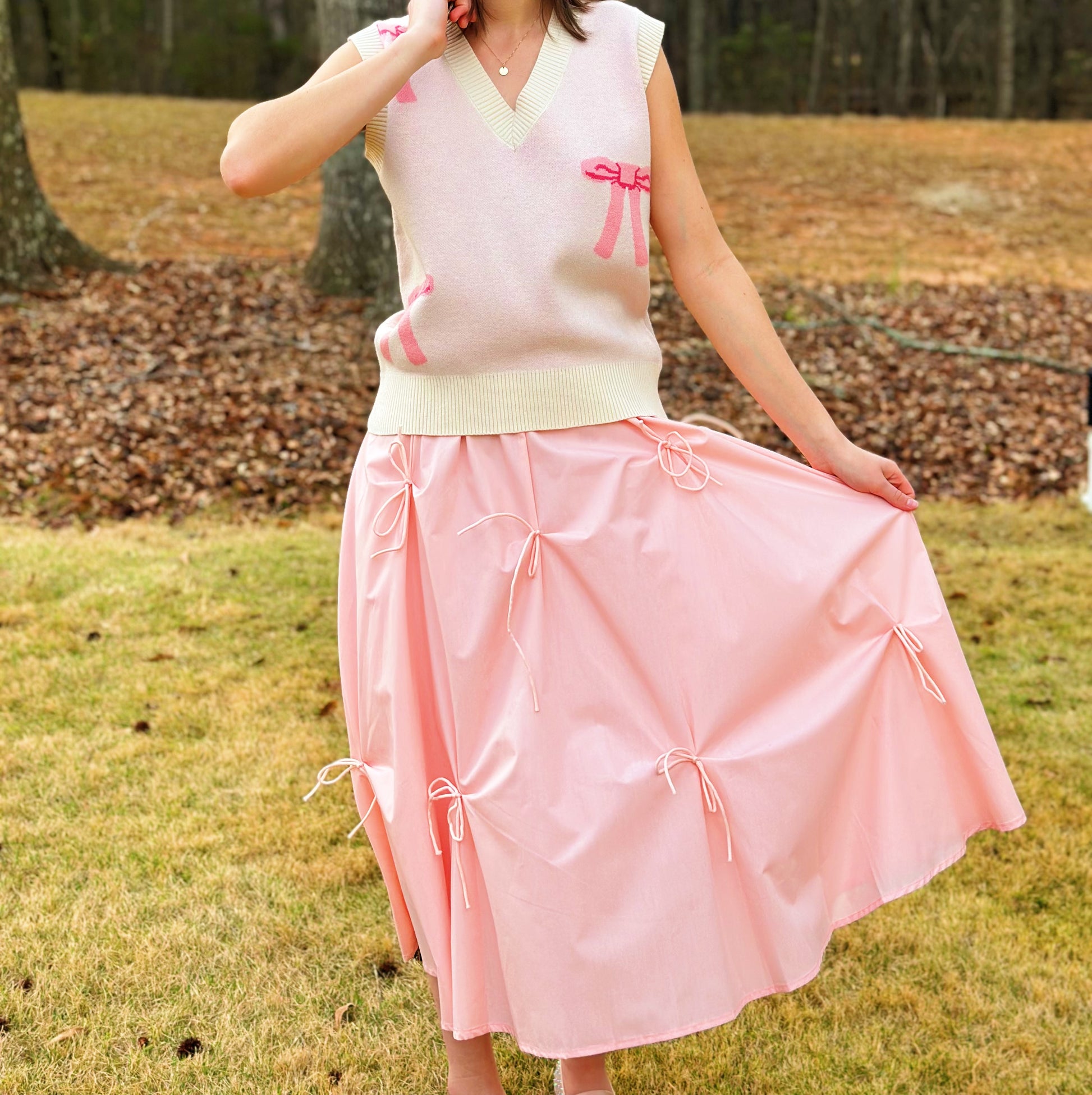 A woman outdoors wearing a sleeveless light pink knit vest with cream trim and pink bow details, paired with a light pink skirt featuring gathered tie details. The background shows grass, fallen leaves, and trees.