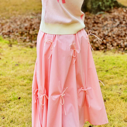 A pink pleated skirt with multiple tie details, worn by a model standing outdoors on a grassy lawn with fallen leaves in the background.