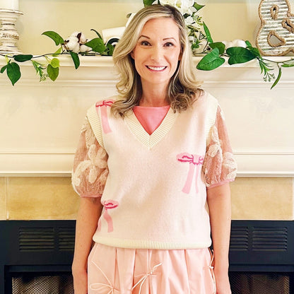 A woman stands in front of a fireplace wearing a light pink knit vest with puff sleeves and matching pink skirt.