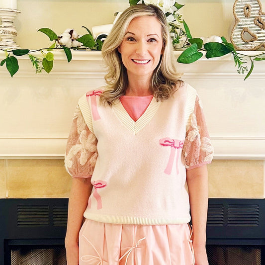 A woman stands in front of a fireplace wearing a light pink knit vest with puff sleeves and matching pink skirt.