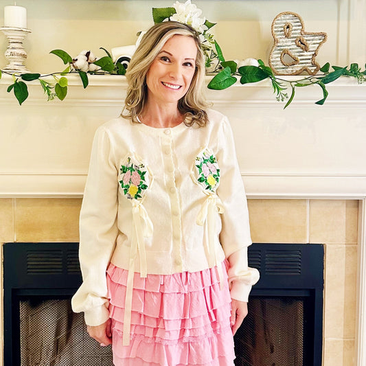 A woman stands in front of a fireplace wearing a cream button-up cardigan with green and yellow floral embroidery, a pink tiered ruffle mini skirt, and white western boots.