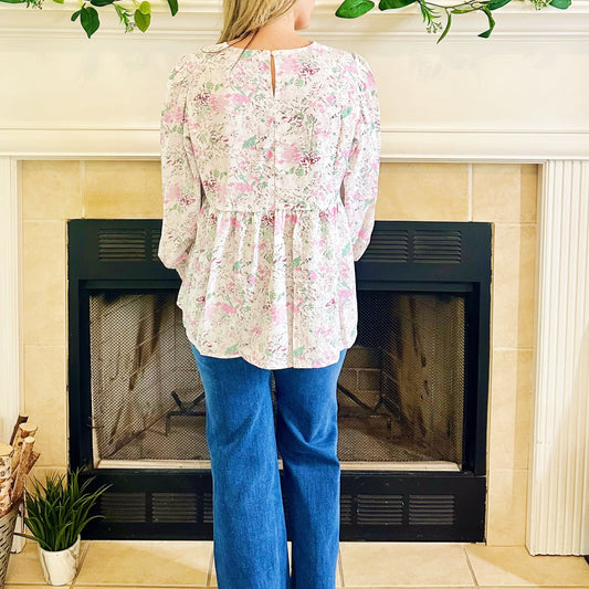 Woman standing in a living room with a fireplace, wearing a floral blouse and blue jeans.