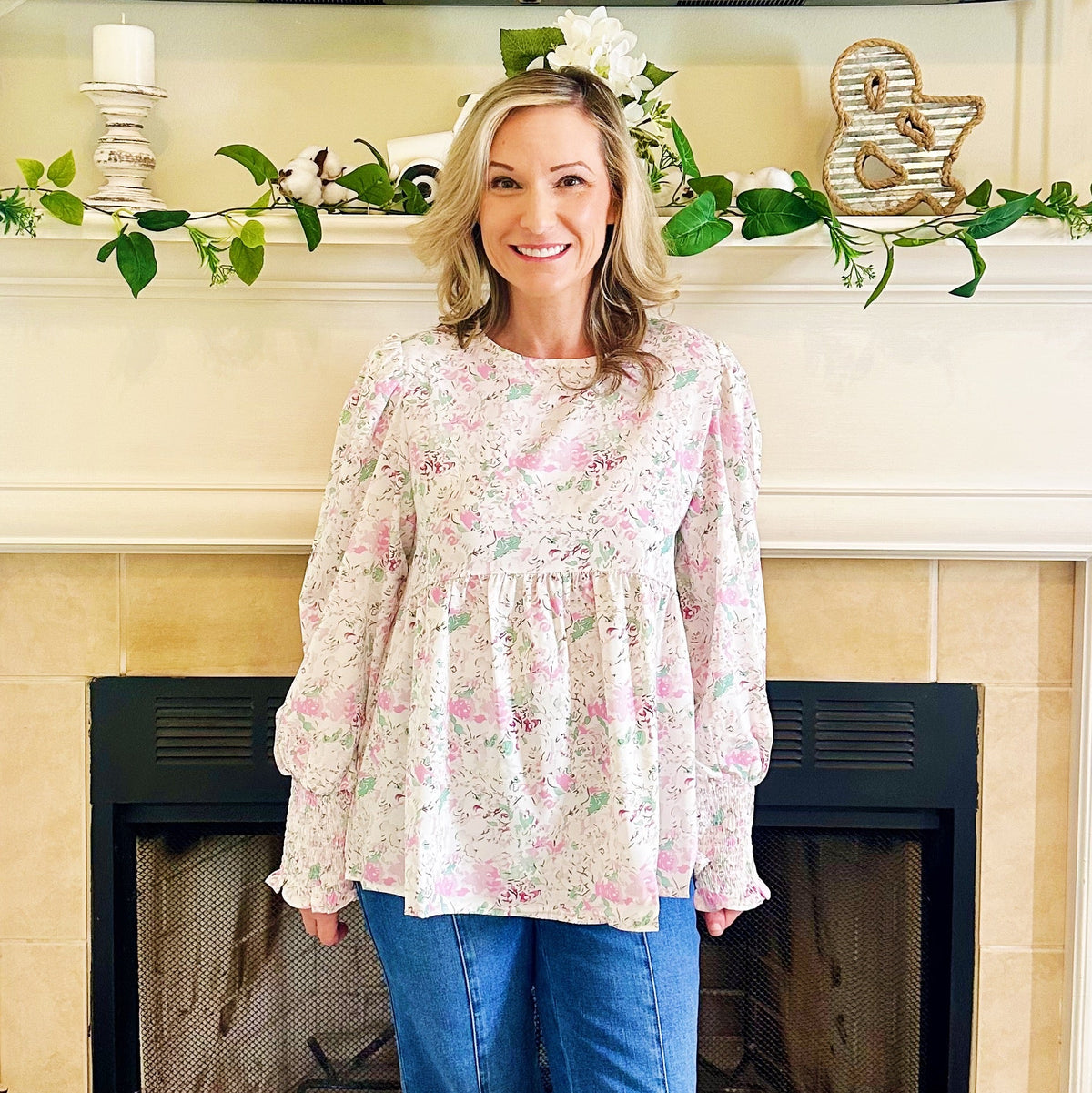 Woman standing in a living room with a fireplace, wearing a floral blouse and blue jeans.