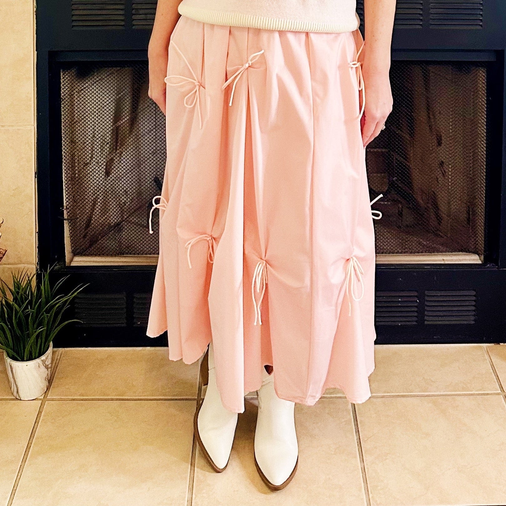 A woman stands in front of a fireplace wearing a light pink knit sweater vest with lace puff sleeves, a pink top underneath, a long light pink tiered skirt with bow details, and white pointed boots.