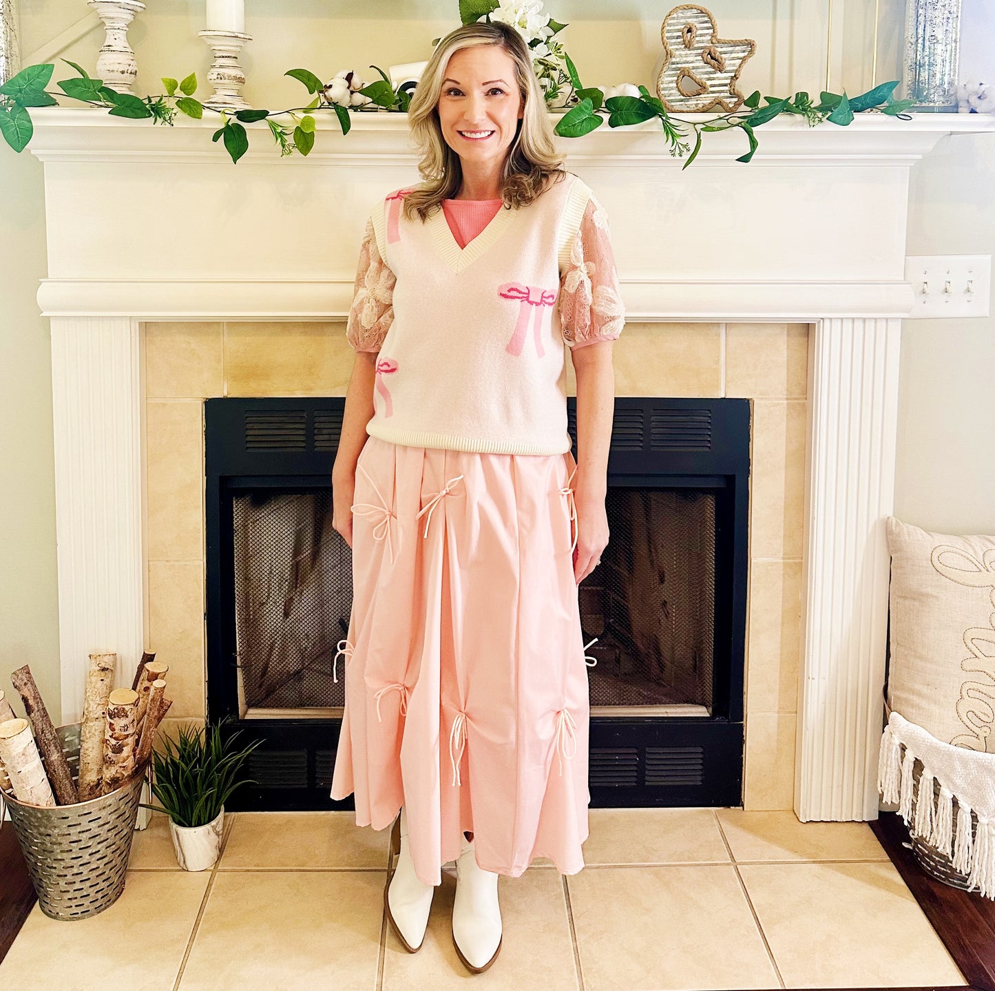 A woman stands in front of a fireplace wearing a light pink knit sweater vest with lace puff sleeves, a pink top underneath, a long light pink tiered skirt with bow details, and white pointed boots.