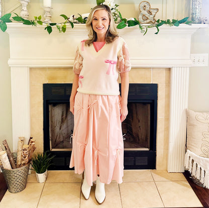 A woman stands in front of a fireplace wearing a light pink knit sweater vest with lace puff sleeves, a pink top underneath, a long light pink tiered skirt with bow details, and white pointed boots.