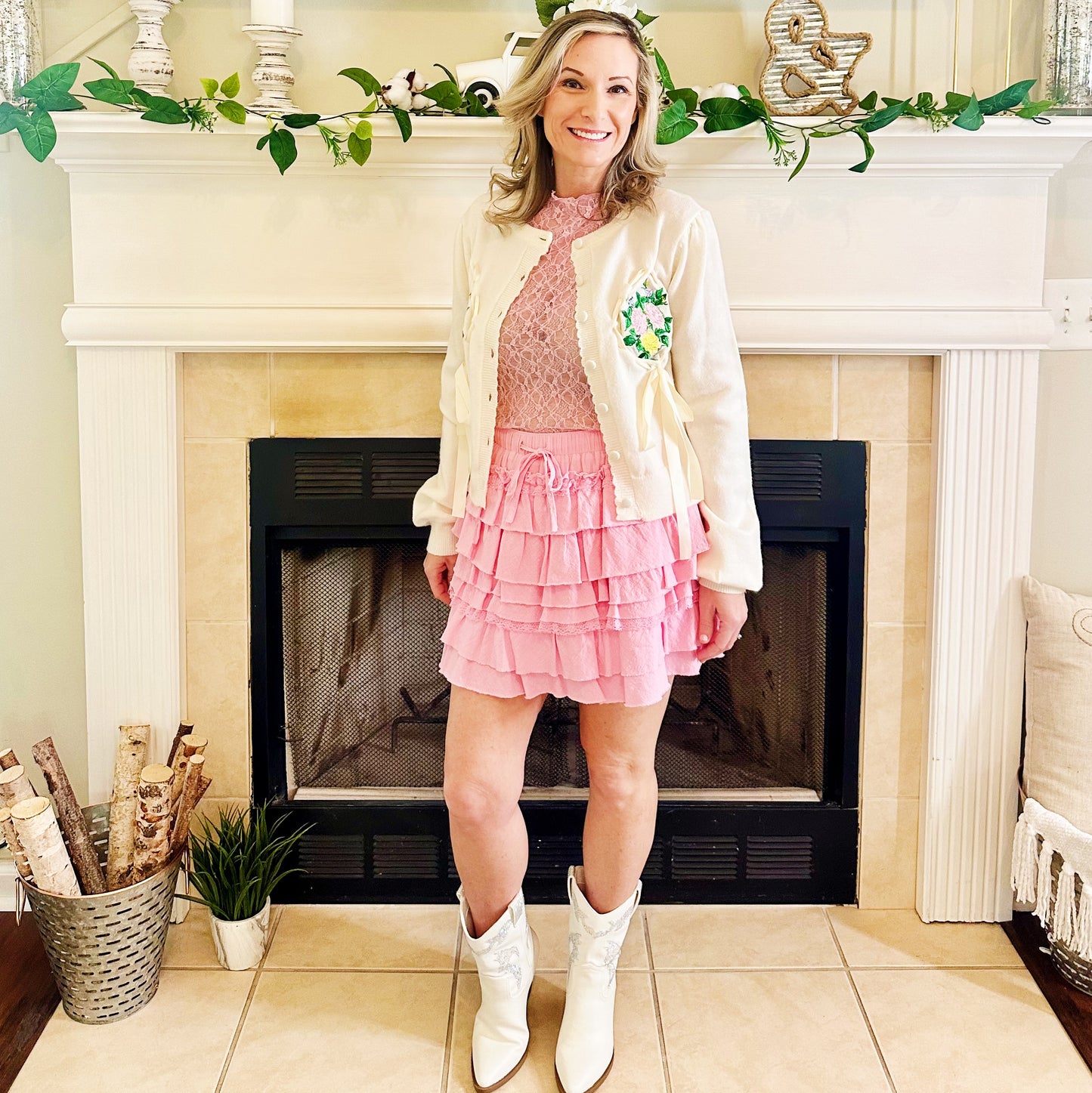 A woman stands in front of a fireplace wearing a pink ruffled mini skirt, a pink lace top, a cream knit cardigan with a green floral patch, and white cowboy boots.