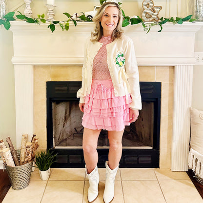 A woman stands in front of a fireplace wearing a pink ruffled mini skirt, a pink lace top, a cream knit cardigan with a green floral patch, and white cowboy boots.