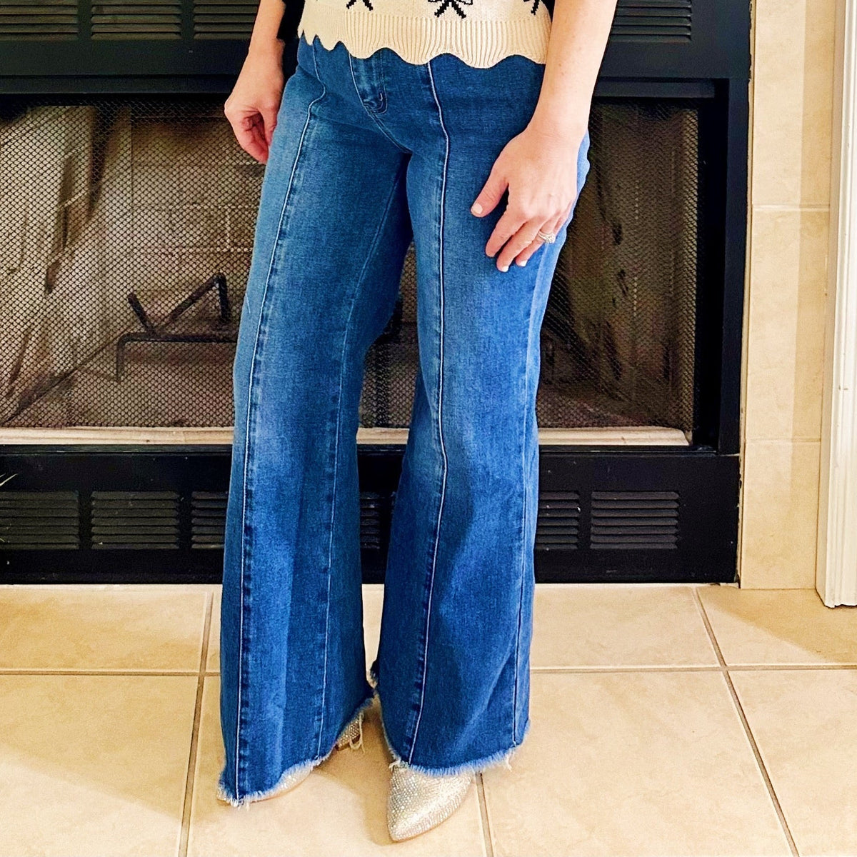 A woman stands in front of a fireplace wearing a cream short-sleeve top with a black bow pattern and blue wide-leg jeans.