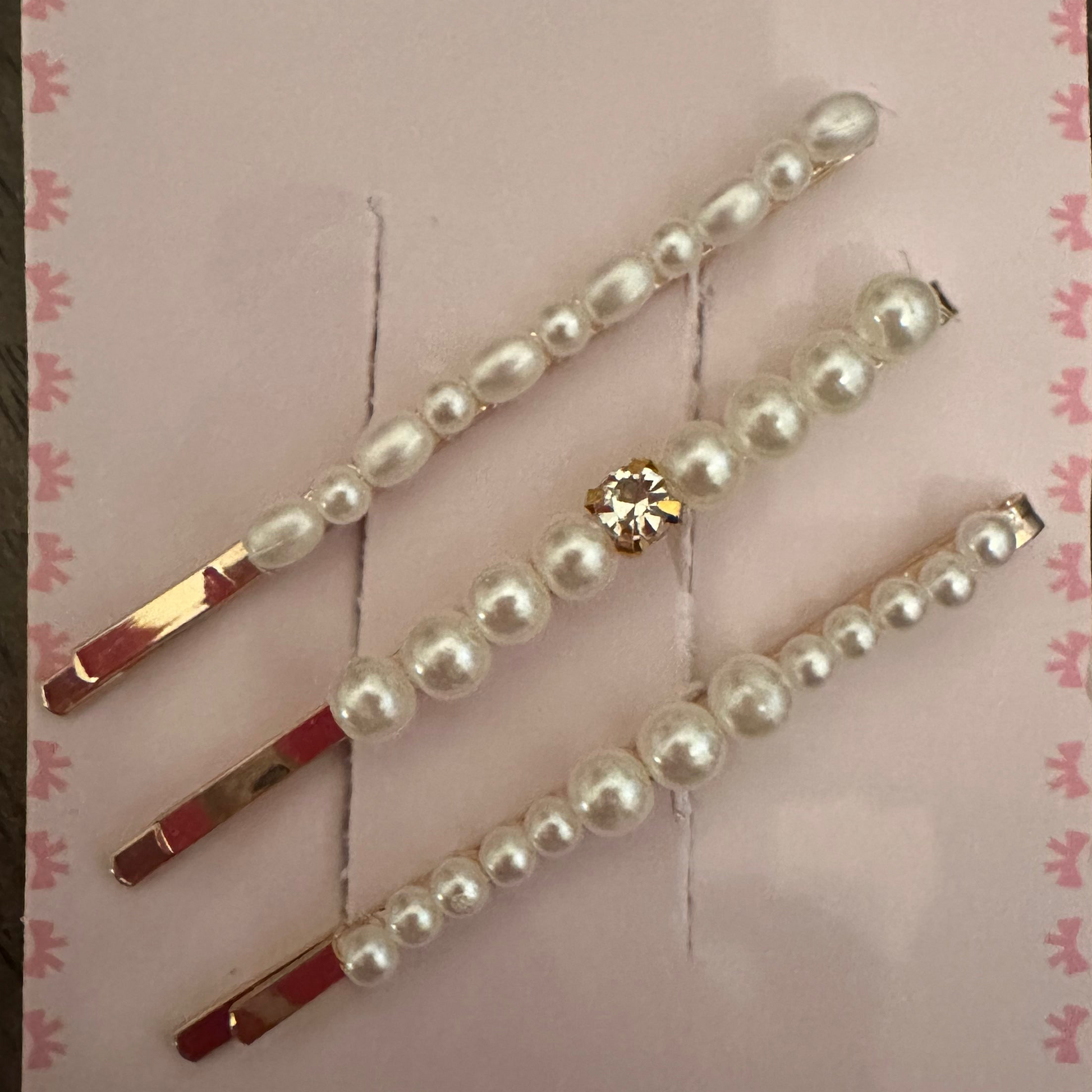 Three pearl hair pins with gold-tone metal bases are displayed on a pink card with a floral border, set against a dark wood background.