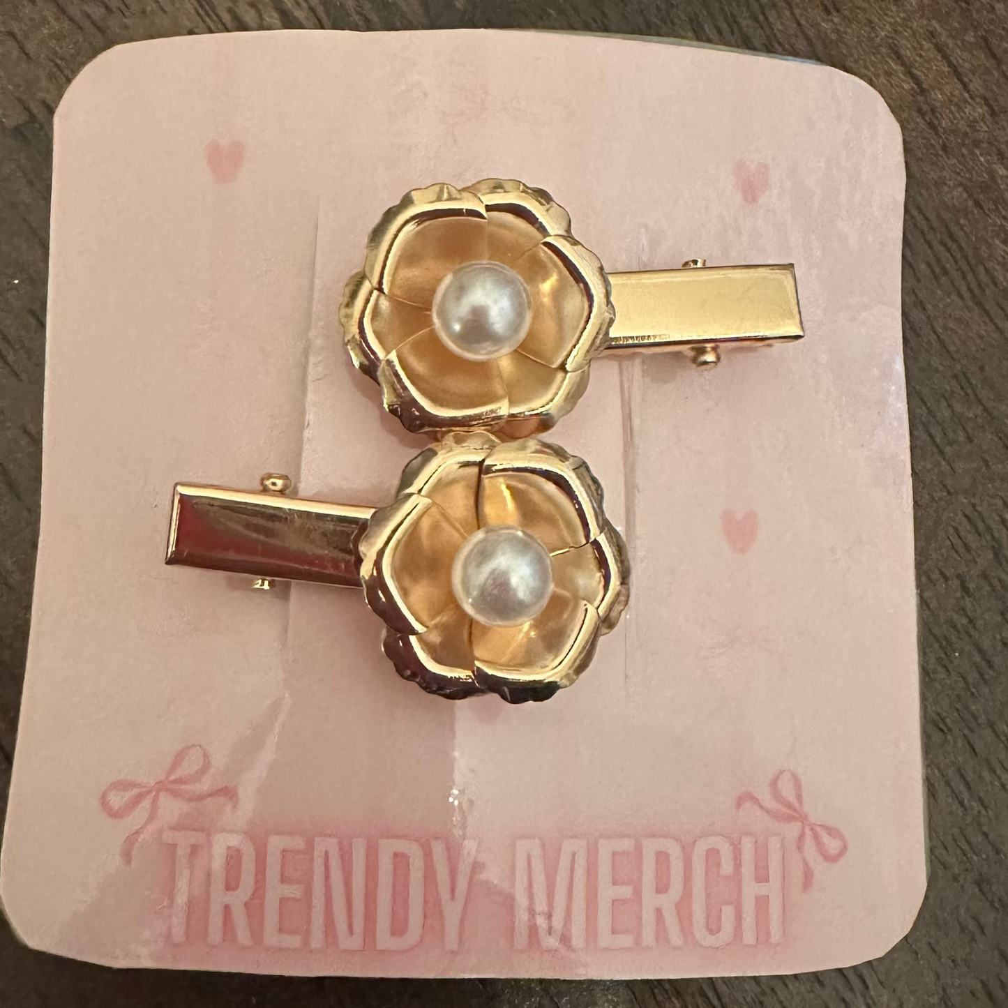 A pair of gold-tone hair clips with pearl centers, presented on a pink card labeled 'TRENDY MERCH' on a dark wooden table.