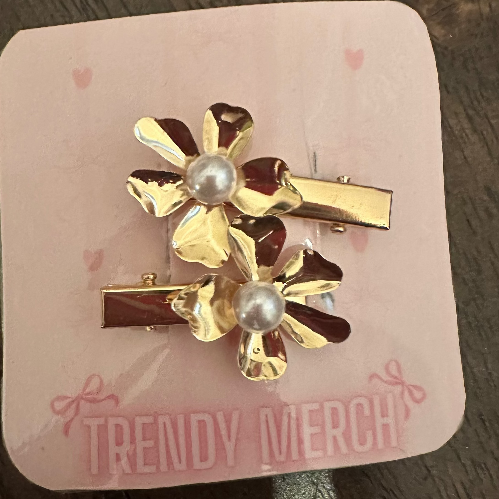 Two gold flower-shaped hair clips with pearl centers, displayed on a pink card labeled 'TRENDY MERCH' on a dark wooden surface.