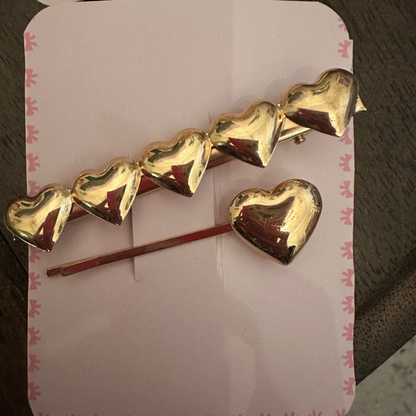 A gold heart-themed hair accessory set consisting of a barrette with five connected heart charms and a separate heart pin, displayed on a pink card with a bow pattern, set against a dark wooden background.