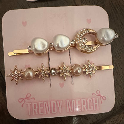 Two gold hair clips with pearl and crystal details, displayed on a pink card labeled 'TRENDY MERCH' on a dark wooden table.