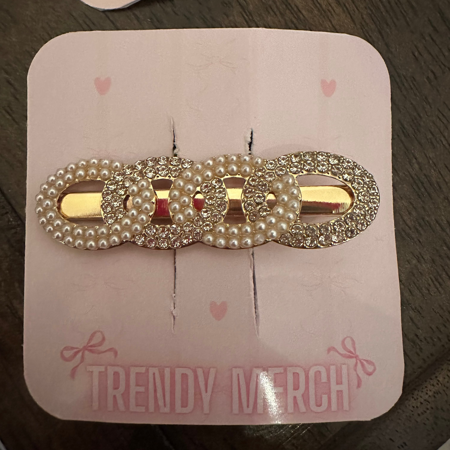 A gold-tone hair claw clip embellished with faux pearls and rhinestones, displayed on a pink card labeled 'TRENDY MERCH' on a dark wooden surface.