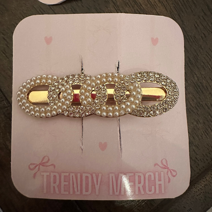A gold-tone hair claw clip embellished with faux pearls and rhinestones, displayed on a pink card labeled 'TRENDY MERCH' on a dark wooden surface.