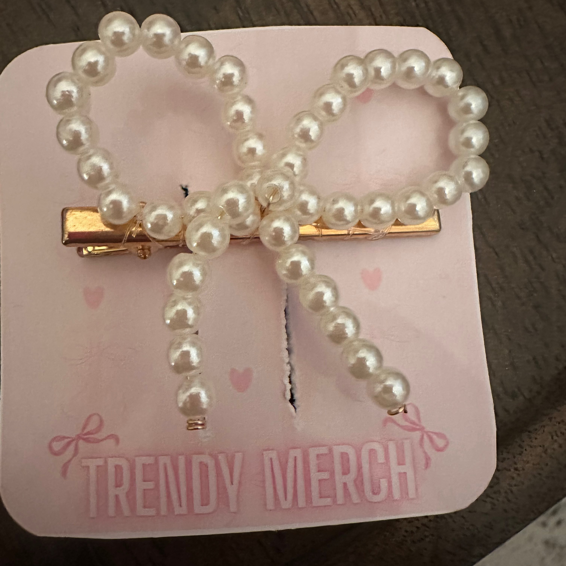 A pearl-bead bow-shaped hair clip with a gold-tone metal barrette base, displayed on a pink 'TRENDY MERCH' backing card on a dark wooden surface.