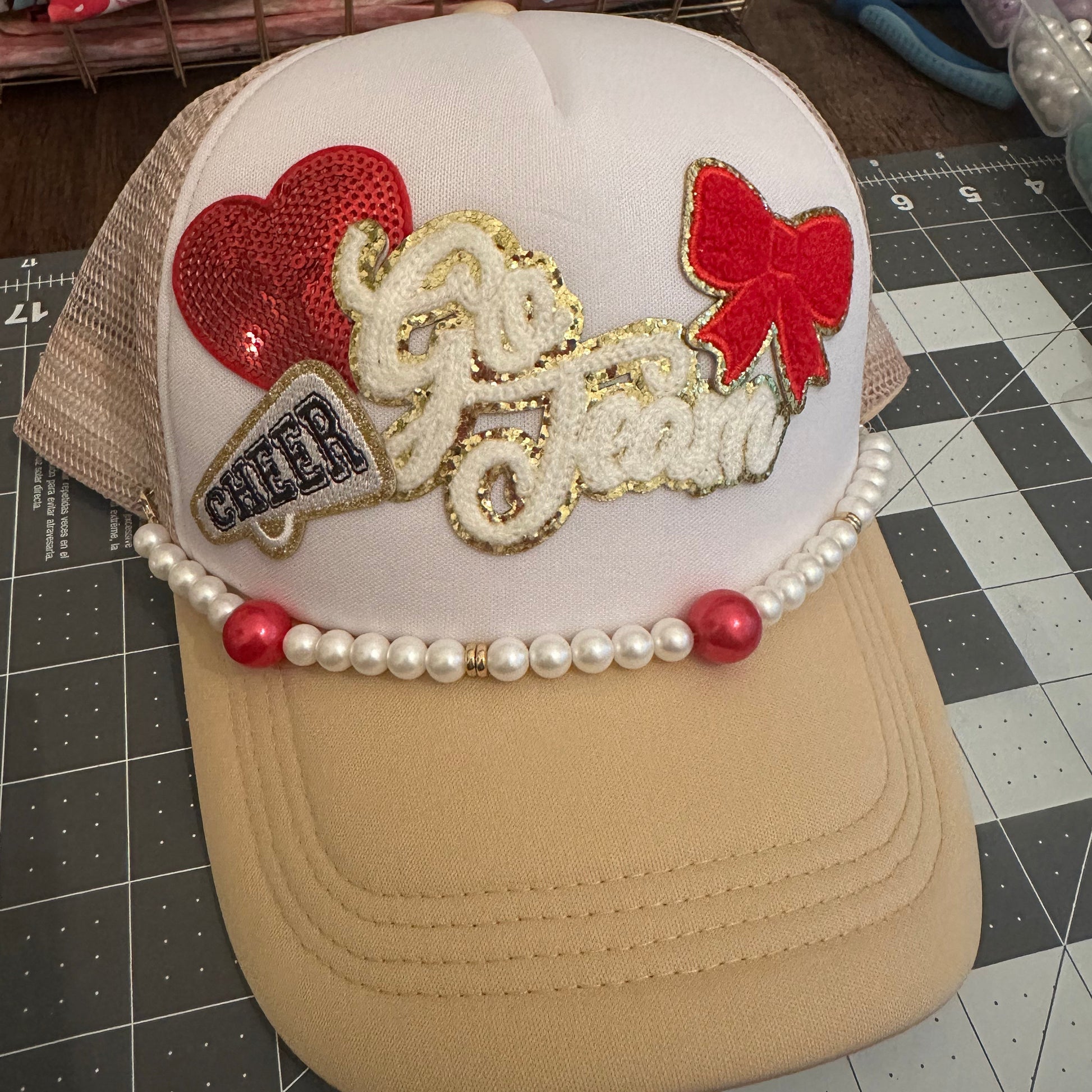 A custom trucker-style cap with a white front panel, beige mesh sides, and a beige brim. The front is decorated with a red sequin heart, a red bow, a gold glitter script reading 'Go Jean', a small 'CHIC' patch, and a pearl bead trim with red beads.