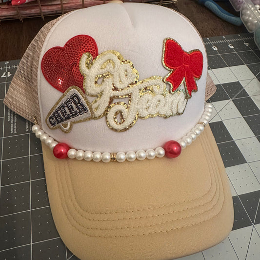 A custom trucker-style cap with a white front panel, beige mesh sides, and a beige brim. The front is decorated with a red sequin heart, a red bow, a gold glitter script reading 'Go Jean', a small 'CHIC' patch, and a pearl bead trim with red beads.