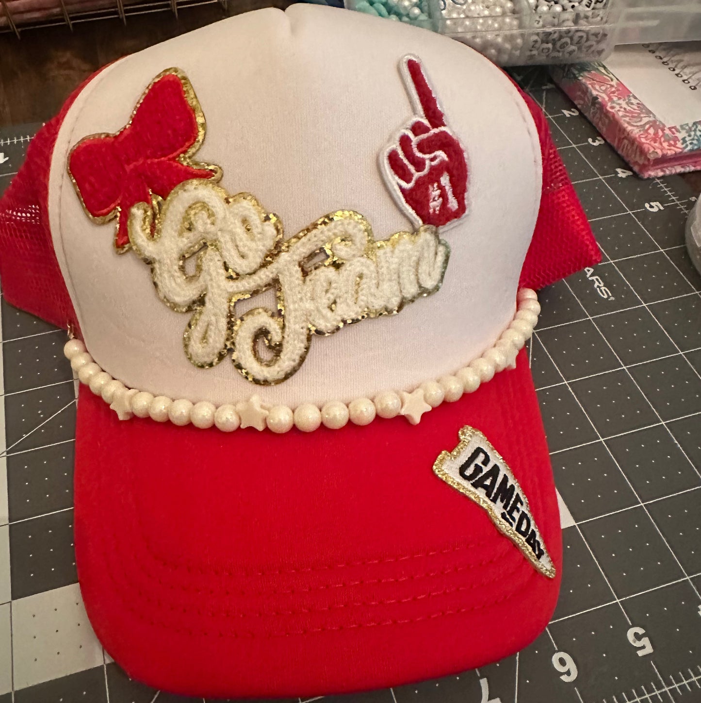 A red and white trucker hat with custom embroidery. The front panel is white with a red bow, a red hand gesture patch, and 'Go Team' in white script with gold glitter trim.