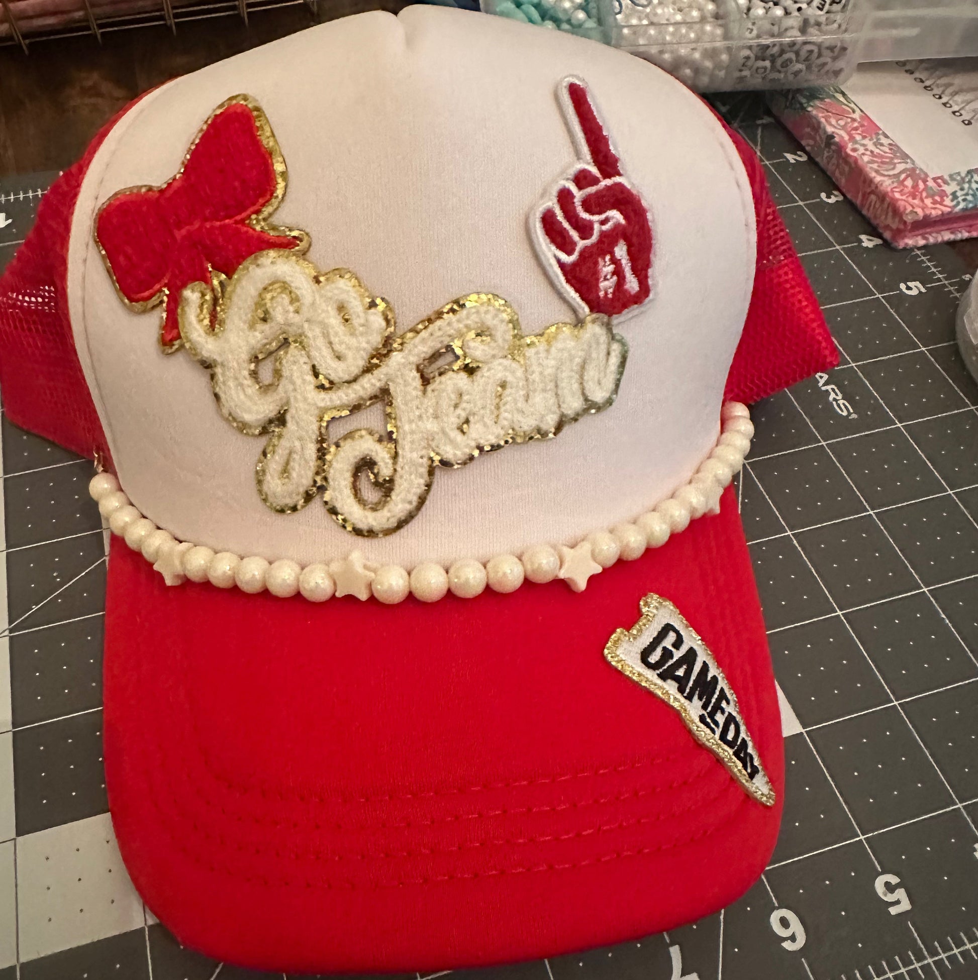A red and white trucker hat with custom embroidery. The front panel is white with a red bow, a red hand gesture patch, and 'Go Team' in white script with gold glitter trim.