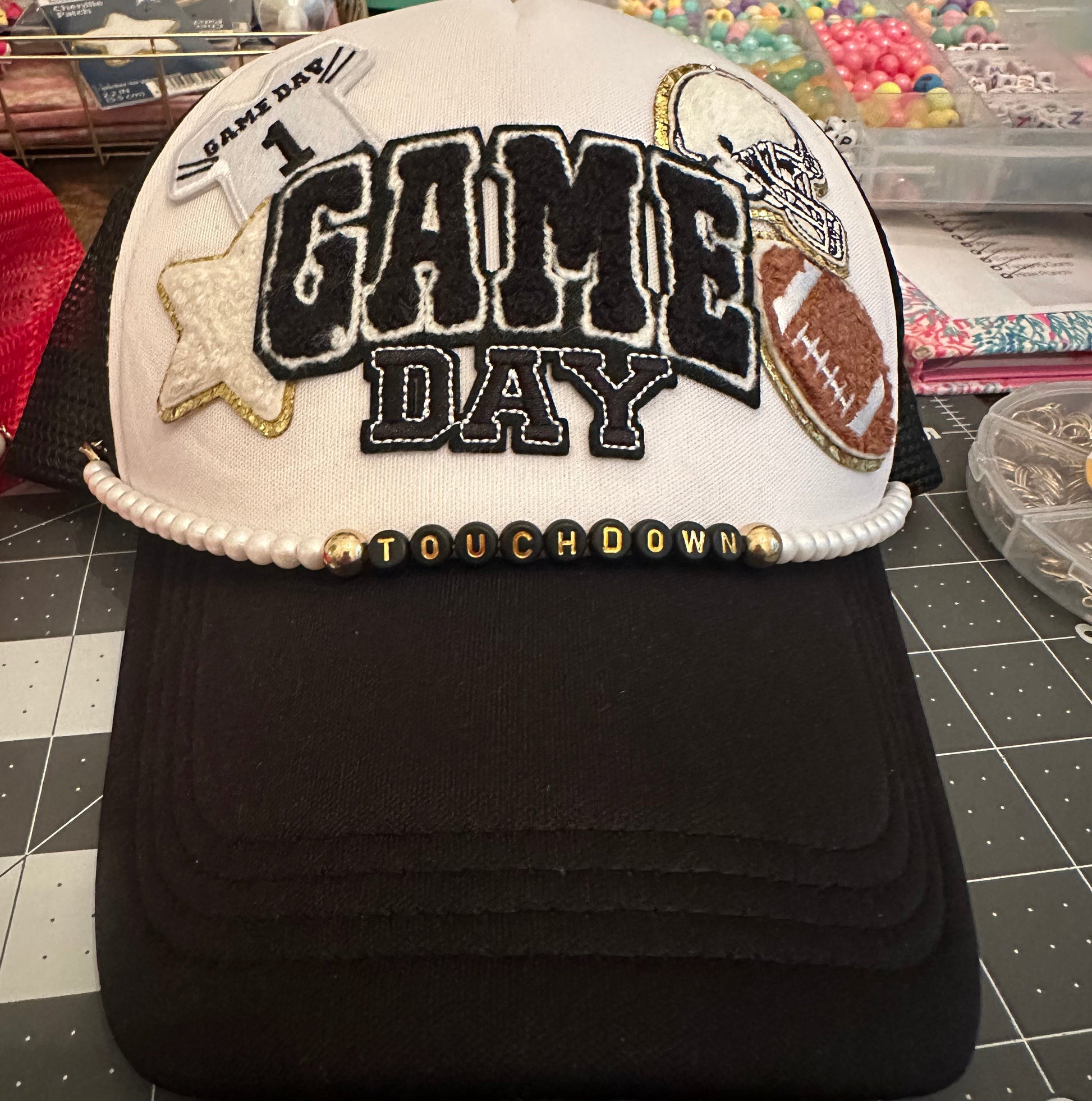 A custom white and black trucker-style cap featuring 'GAME DAY' embroidered on the front, a gold star patch, a football patch, and a beaded 'TOUCHDOWN' trim, displayed on a cutting mat with craft supplies in the background.