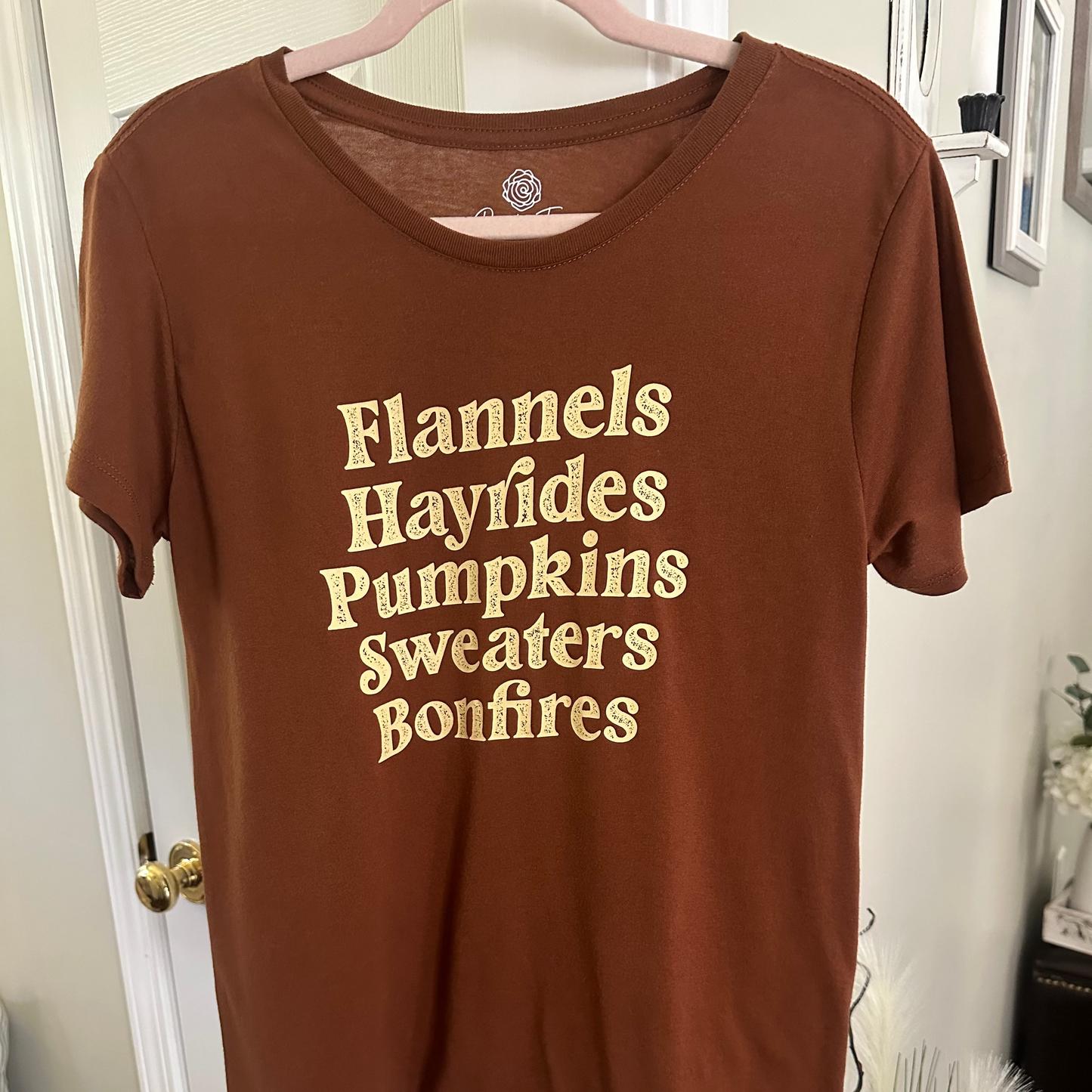 A brown short-sleeve t-shirt hanging on a pink hanger against a white door, featuring the words 'Flannels Hayfides Pumpkins Sweaters Bonfires' printed in a beige serif font on the front.