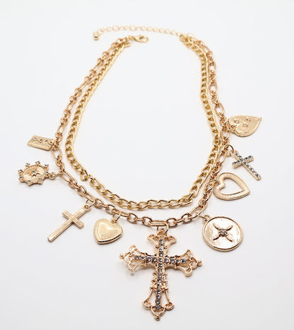 Charm Necklace. This beautifully layered gold-tone necklace features an enchanting collection of delicate charms, including graceful hearts, intricate crosses, and classic coin pendants. The centerpiece, a stunningly detailed cross.