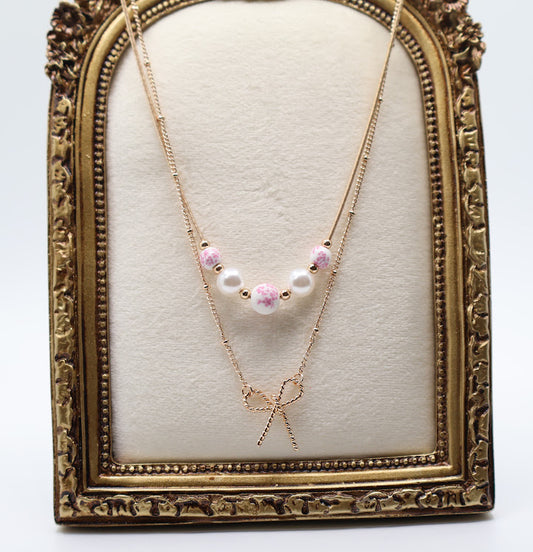 Bow & Bead Layered Necklace. This layered design features a dainty gold-tone bow pendant on one chain and pocelain white beads with pink floral print. 