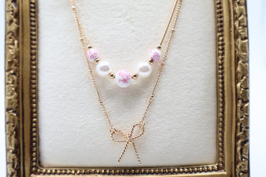  Bow & Bead Layered Necklace. This layered design features a dainty gold-tone bow pendant on one chain and pocelain white beads with pink floral print. 
