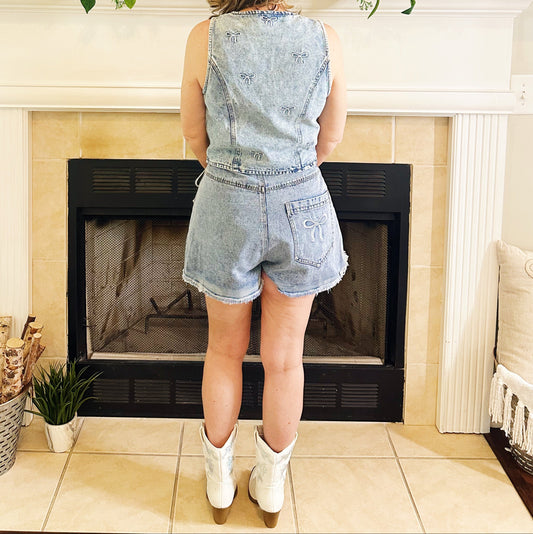 A woman stands with her back to the camera in front of a fireplace, wearing a light blue denim sleeveless romper with matching denim shorts and white cowboy boots.