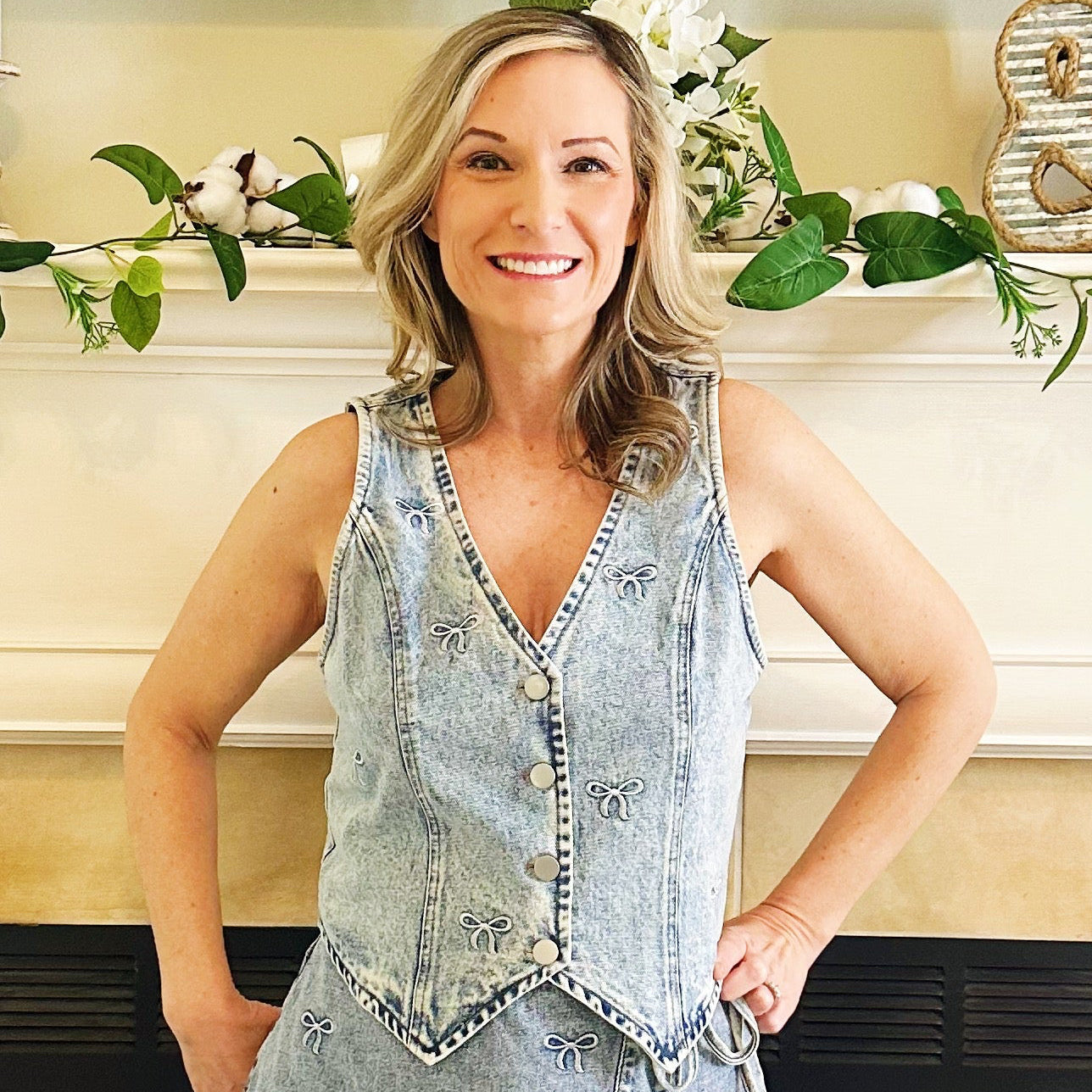 A woman stands in front of a fireplace wearing a sleeveless light-wash denim mini dress with a frayed hem and white cowboy boots.