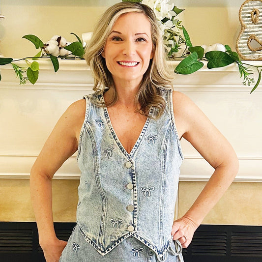 A woman stands in front of a fireplace wearing a sleeveless light-wash denim mini dress with a frayed hem and white cowboy boots.