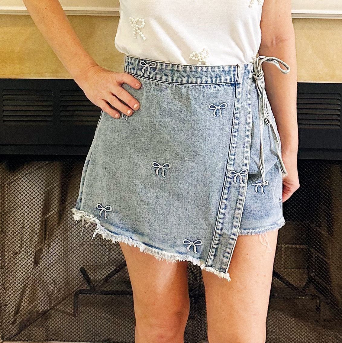 A person wearing a light blue denim wrap mini skirt with embroidered bow details and a frayed hem, paired with a white top, standing in front of a fireplace.