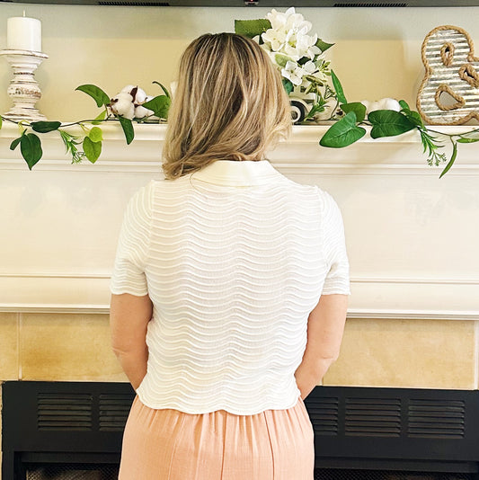 A woman viewed from behind wearing a short-sleeve textured cream knit top and a peach skirt, standing in front of a fireplace mantel decorated with greenery and white flowers.