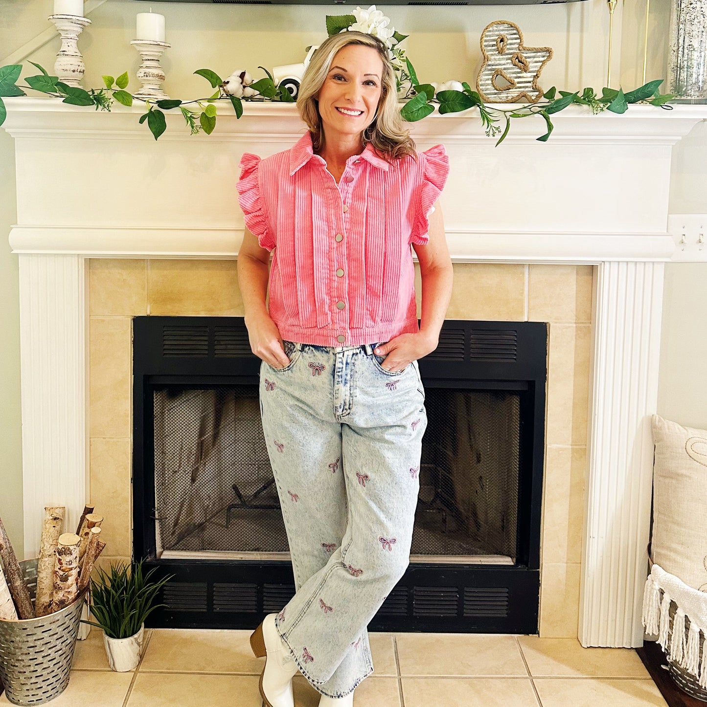 A woman stands in front of a fireplace wearing a pink ruffled sleeveless button-up top, light-wash high-waisted jeans with small floral embroidery, and white cowboy boots.