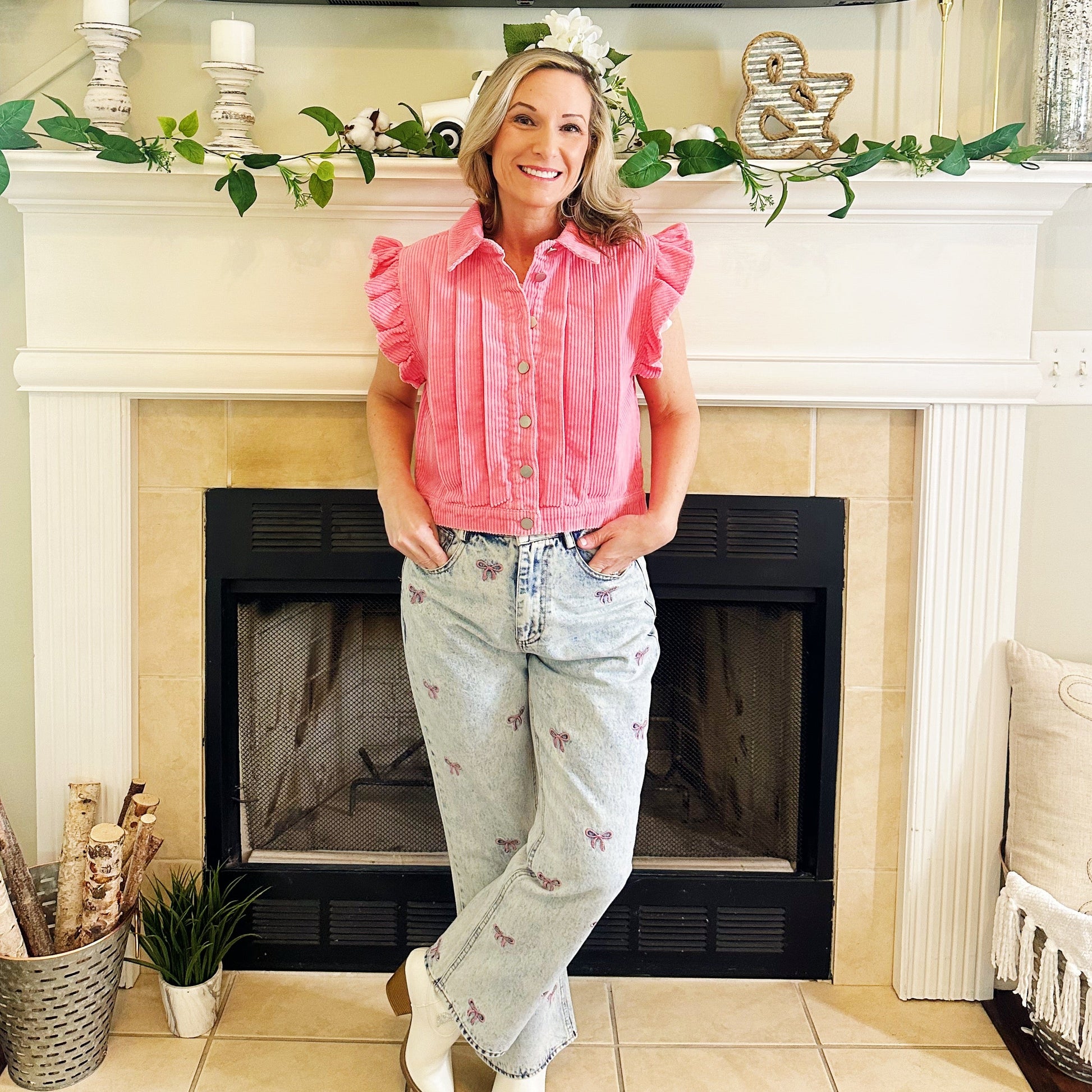 A woman stands in front of a fireplace wearing a pink ruffled sleeveless button-up top, light-wash high-waisted jeans with small floral embroidery, and white cowboy boots.