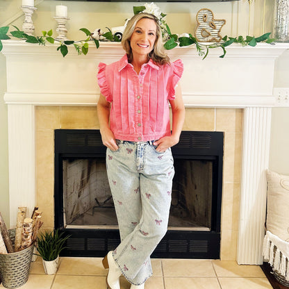 A woman stands in front of a fireplace wearing a pink ruffled sleeveless button-up top, light-wash high-waisted jeans with small floral embroidery, and white cowboy boots.